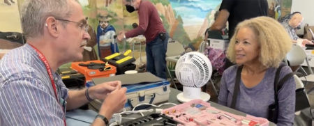 Man with glasses and a lanyard explains a tool kit to a smiling woman at a community workshop, with a pink tool case and a small fan on the table nearby.