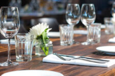 Table set for dinner with wine glasses, water glasses, plates, and a small white flower centerpiece.