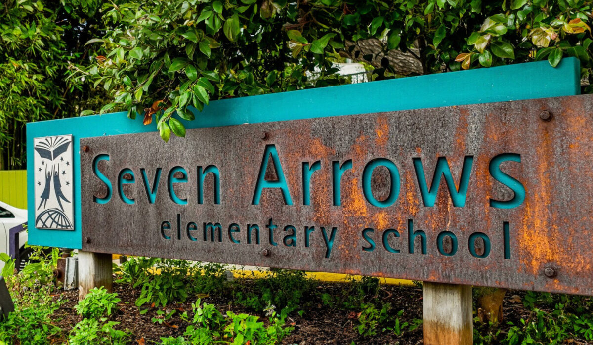 Seven Arrows Elementary School sign on a rusted metal panel with a teal border and greenery around it.