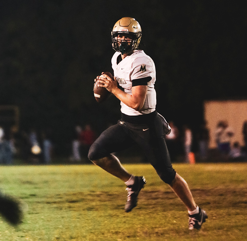 Football quarterback in white jersey and gold helmet, mid-run and preparing to throw a pass on a nighttime field