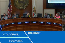 City Council chamber with members around a curved dais; flags and sunflowers on desks; blue 'Public Input' banner with date 09/22/2026 in front.