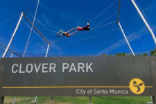 Sign for Clover Park, City of Santa Monica, with an outdoor high ropes course in the background and two people walking on a cable above the sign.