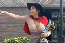 Woman in a wide-brimmed hat points to the left while holding a skateboard outdoors.
