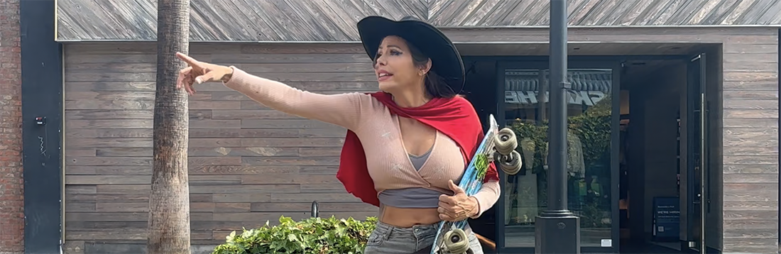 Woman in a wide-brimmed hat points to the left while holding a skateboard outdoors.