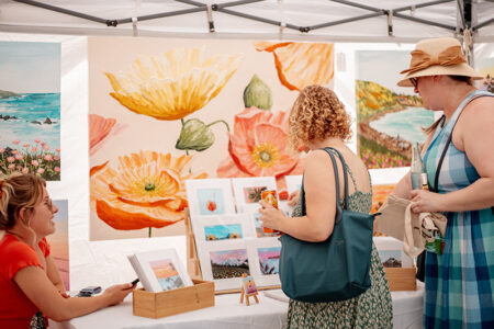 Outdoor art market stall with large yellow and orange flower paintings; three women browse framed prints.