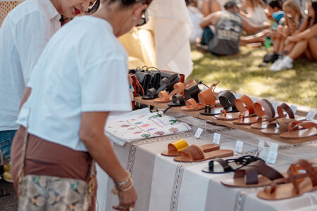 Shoppers browse a table of leather sandals at an outdoor market stall on a sunny day.