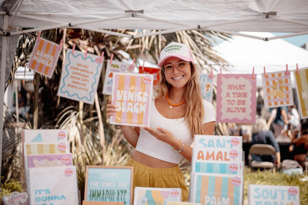 Smiling woman in a white crop top and mustard skirt under an outdoor market canopy, holding a pastel striped print that says 'VENICE BEACH' with more colorful art prints displayed around her.