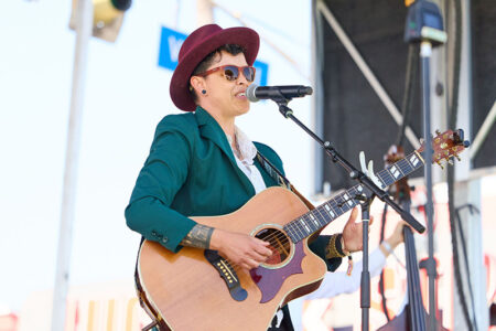 Musician performing outdoors with an acoustic guitar and microphone, wearing a burgundy hat, sunglasses, and a teal blazer on stage.