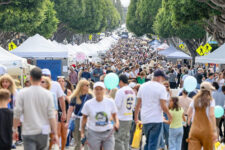 Crowded street festival with rows of white vendor tents and people walking along a tree-lined avenue.