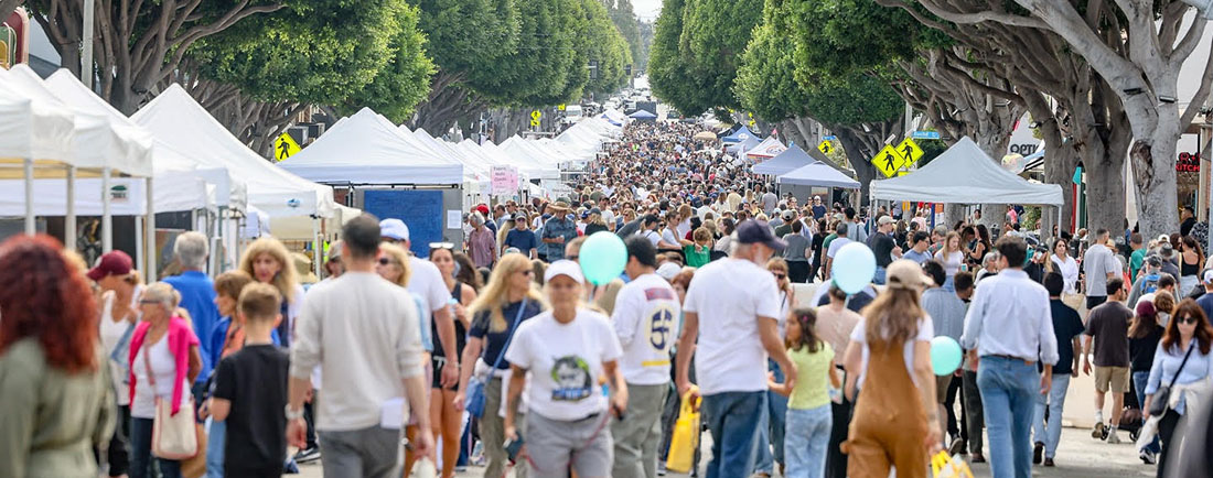 Crowded street festival with rows of white vendor tents and people walking along a tree-lined avenue.