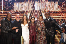 Six people pose for a group photo on a brightly lit talent show stage with a fireworks backdrop and a large 'NENE ROYAL' sign above them, suggesting judges or hosts.