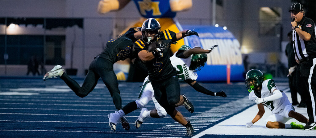 Black-uniformed player carries the ball and charges forward as defenders reach in on a blue turf field during a football play.