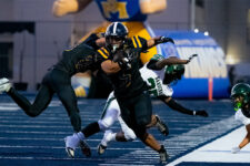 Black-uniformed player carries the ball and charges forward as defenders reach in on a blue turf field during a football play.