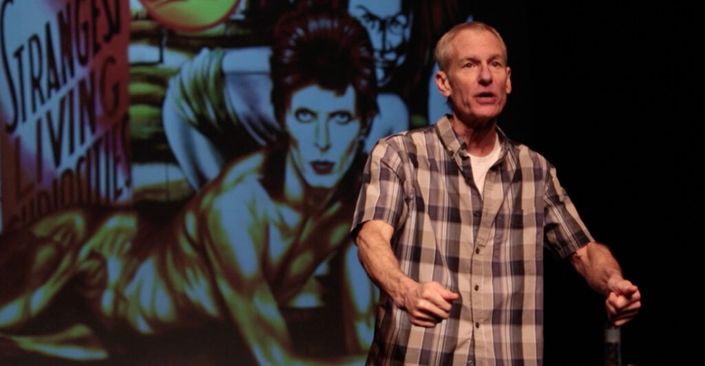 Middle-aged man in a blue plaid shirt giving a talk on a stage, with a large colorful poster or mural featuring a person in the background on the left side of the scene.