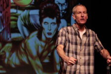 Middle-aged man in a blue plaid shirt giving a talk on a stage, with a large colorful poster or mural featuring a person in the background on the left side of the scene.