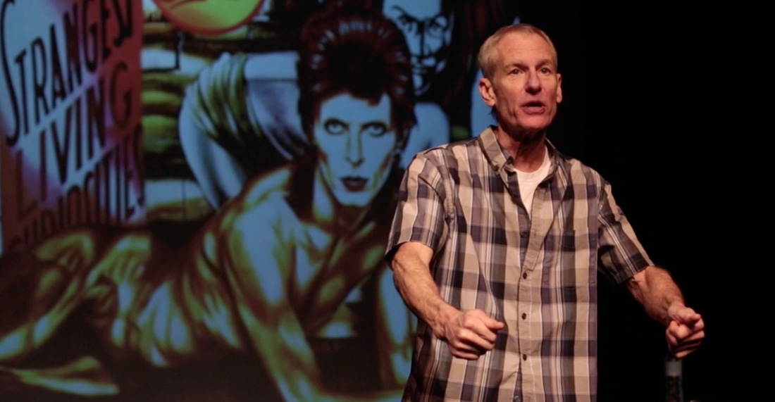 Middle-aged man in a blue plaid shirt giving a talk on a stage, with a large colorful poster or mural featuring a person in the background on the left side of the scene.
