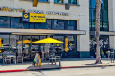California Pizza Kitchen storefront with outdoor seating under yellow umbrellas on a sunny street, with a palm tree and a white car parked nearby.