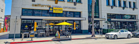 California Pizza Kitchen storefront with outdoor seating under yellow umbrellas on a sunny street, with a palm tree and a white car parked nearby.