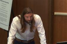 Man with long hair and beard leaning over a podium, speaking into a microphone in a conference room.