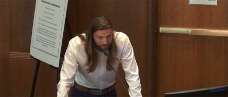 Man with long hair and beard leaning over a podium, speaking into a microphone in a conference room.