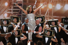 Singer on stage in a silver dress is lifted by dancers wearing retro TV-headed costumes with spotlights behind them.