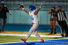 Football player in white and blue uniform celebrates a touchdown in the end zone with arms raised; referee and photographer in background.