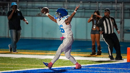 Football player in white and blue uniform celebrates a touchdown in the end zone with arms raised; referee and photographer in background.
