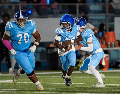 Three football players in light-blue uniforms on a field at night; one carries the ball as two teammates pursue.