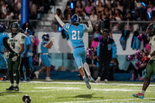 Football player in a blue jersey (#21) jumps mid-air with arms raised in celebration on a football field as teammates and fans watch.