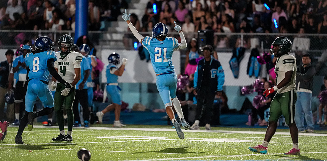 Football player in a blue jersey (#21) jumps mid-air with arms raised in celebration on a football field as teammates and fans watch.