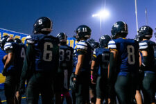 High school football team huddled together on the blue turf under bright stadium lights at dusk.