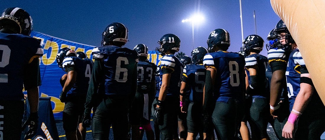 High school football team huddled together on the blue turf under bright stadium lights at dusk.