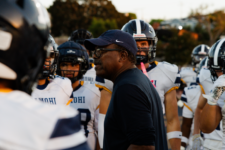 Football coach in a dark shirt and cap speaks to a huddle of players in white jerseys and helmets on the field.