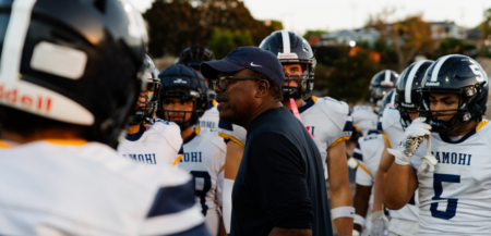 Football coach in a dark shirt and cap speaks to a huddle of players in white jerseys and helmets on the field.