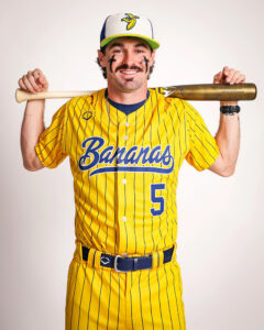 Man in a yellow pinstripe baseball uniform with 'Bananas' script and number 5, holding a bat across his shoulders and smiling on camera.