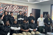 Group of students and instructors posing for a group photo in a barber academy classroom with a large 'Aic Blends Academy' sign behind them.