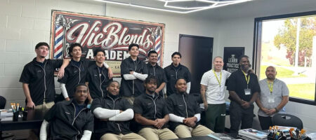 Group of students and instructors posing for a group photo in a barber academy classroom with a large 'Aic Blends Academy' sign behind them.