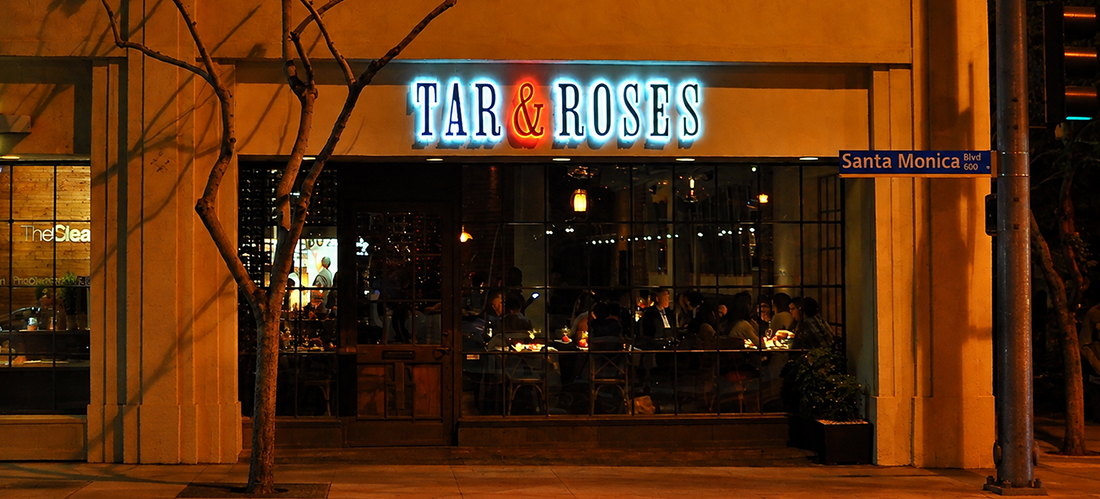 Night view of Tar & Roses restaurant on Santa Monica Blvd, with diners inside through large glass windows.