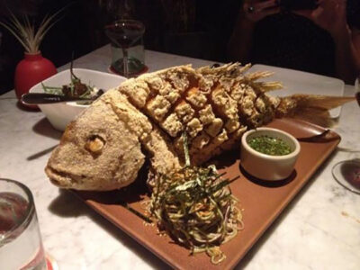 Whole fried fish on a rectangular platter with a small bowl of green herb sauce and a crispy garnish on the side, in a dim restaurant setting.