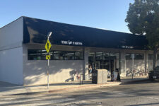Exterior of a small grocery store called The Farms with a black awning, large storefront windows, and a curbside SUV parked on a sunny street.