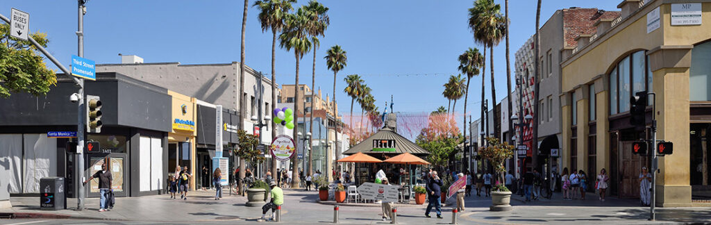 Busy pedestrian street lined with shops and palm trees, with people walking and a gazebo with orange umbrellas at the center.