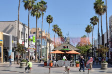 Busy pedestrian street lined with shops and palm trees, with people walking and a gazebo with orange umbrellas at the center.
