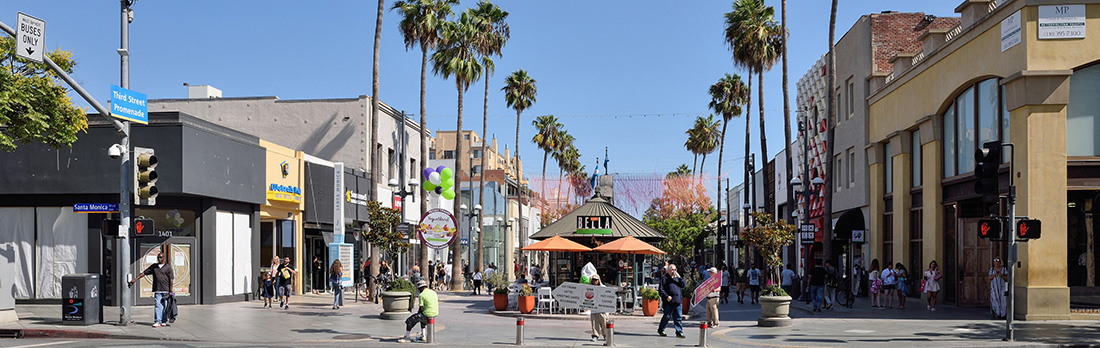 Busy pedestrian street lined with shops and palm trees, with people walking and a gazebo with orange umbrellas at the center.
