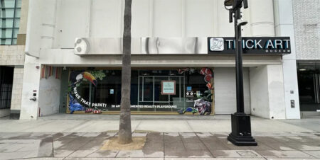 Exterior view of Trick Art Museum storefront with a colorful mural on the glass window and a palm tree in the sidewalk in front.